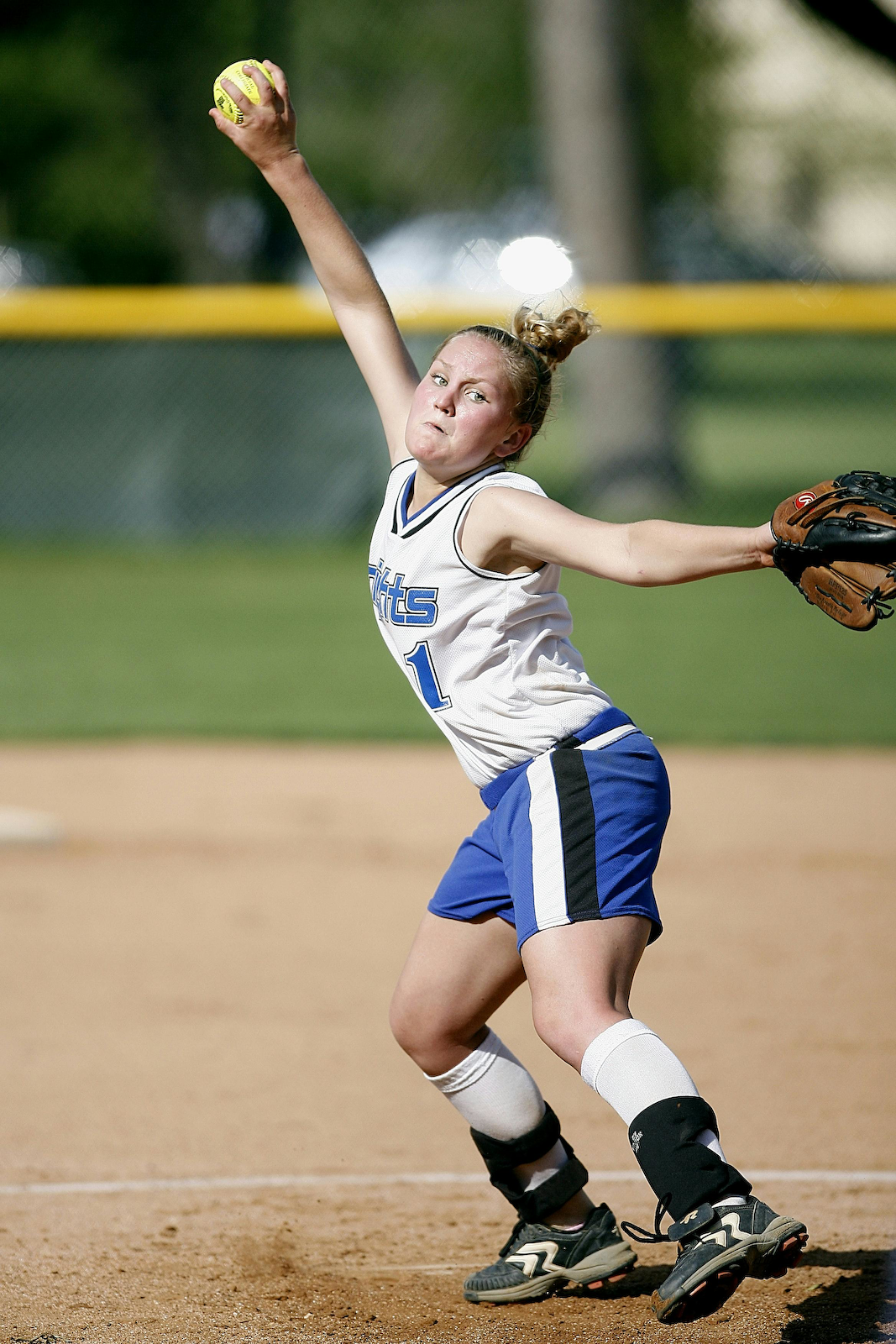 High school softball pitcher in a white and blue uniform mid-windup, throwing a pitch during a game